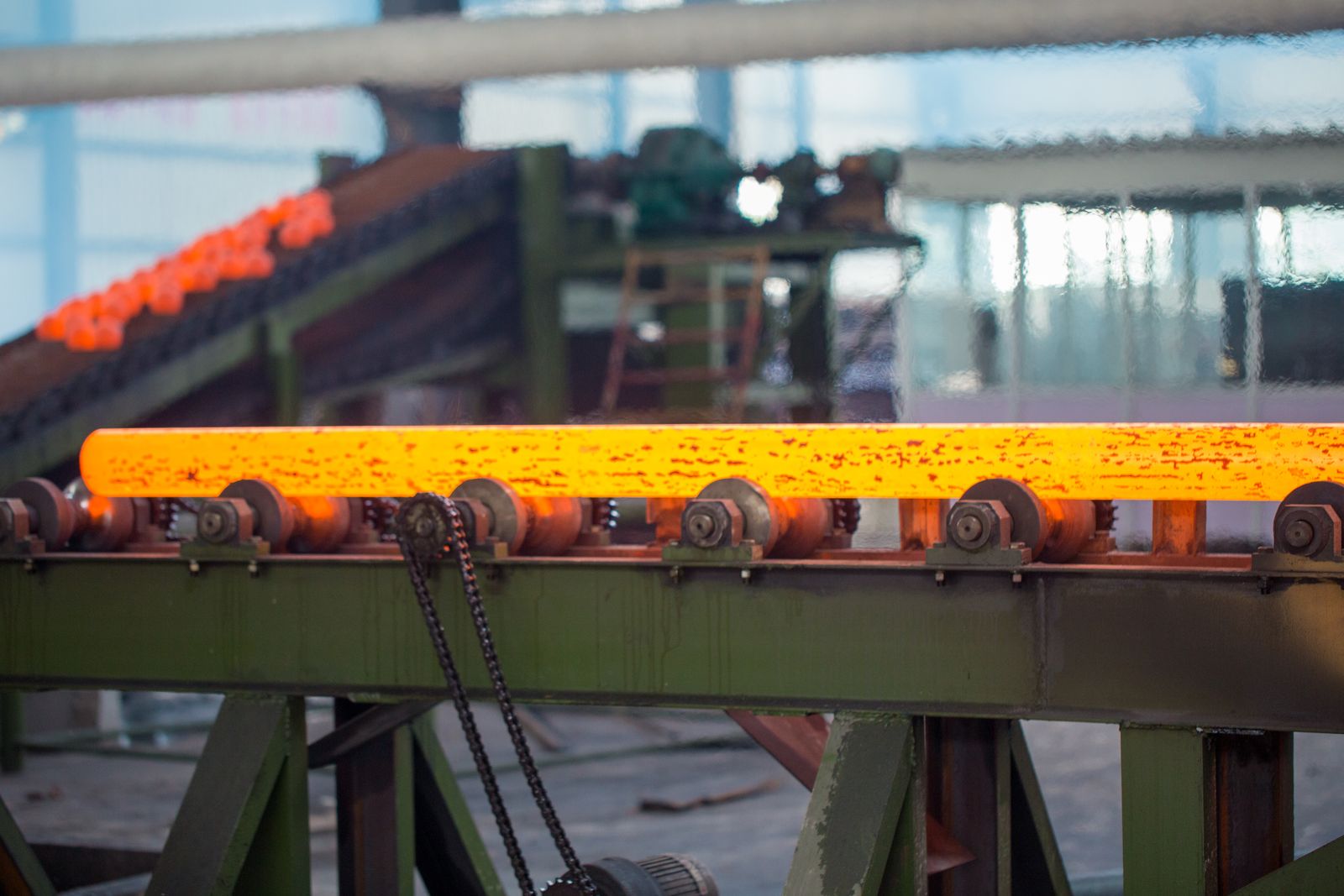 Barra de aço incandescente sobre mesa de roletes, com bolas recém-laminadas ao fundo