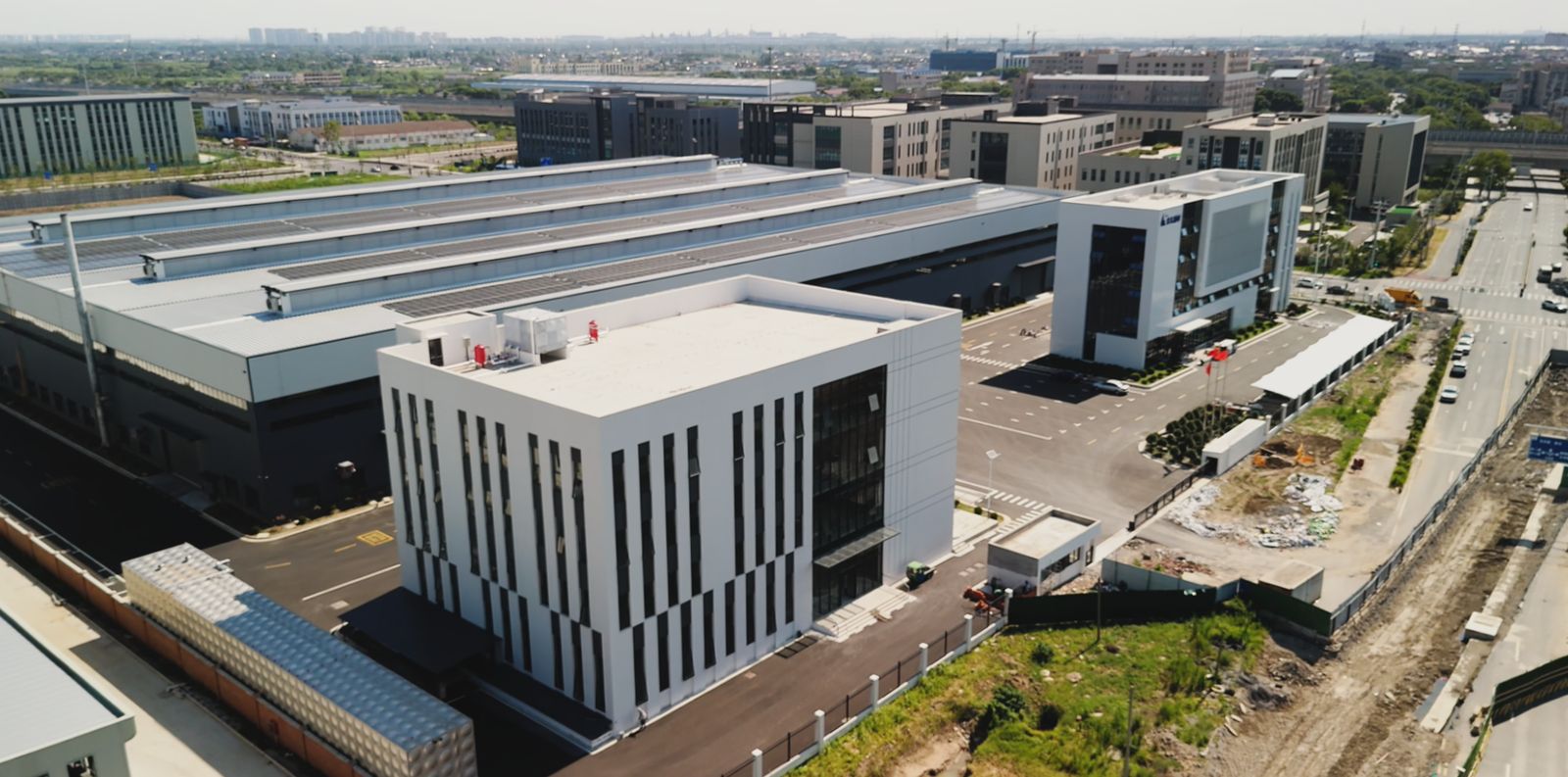 Vista aérea do complexo industrial: prédio administrativo branco e galpões de produção com painéis solares no telhado